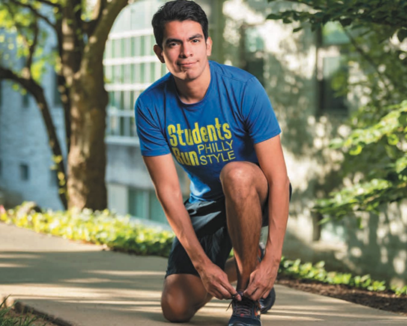 2010 alumnus Luis Gaitan in a light blue T-shirt bending over to tie his shoe laces on a tree-lined street