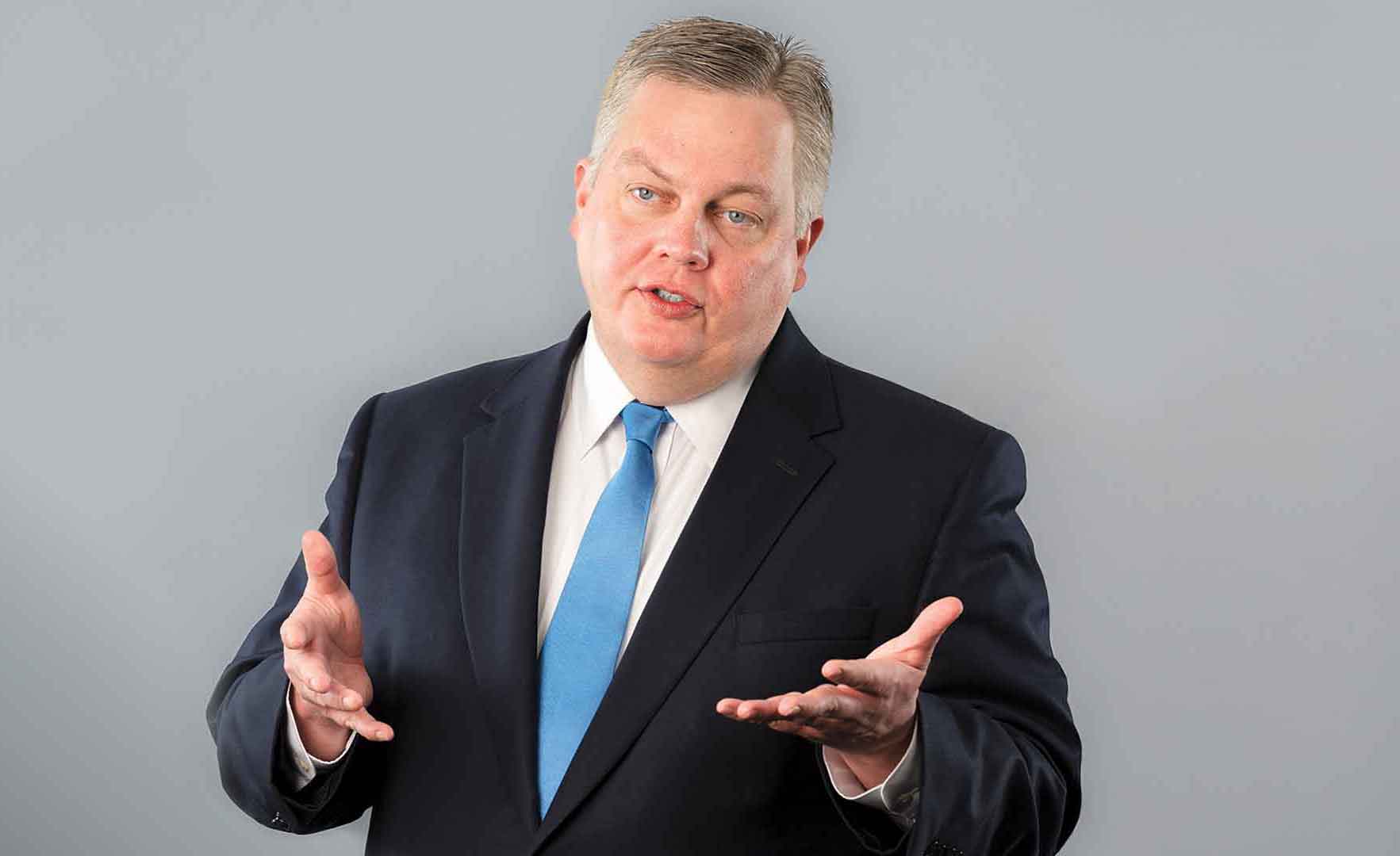 Mike Moreland talking with his hands in a navy suit jacket and blue tie