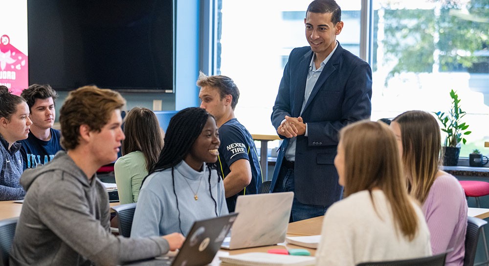 Faculty member standing and talking to group of students seated around a table Faculty member standing and talking to group of students seated around a table