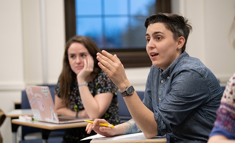 A student speaks in a classroom with another student in the background.