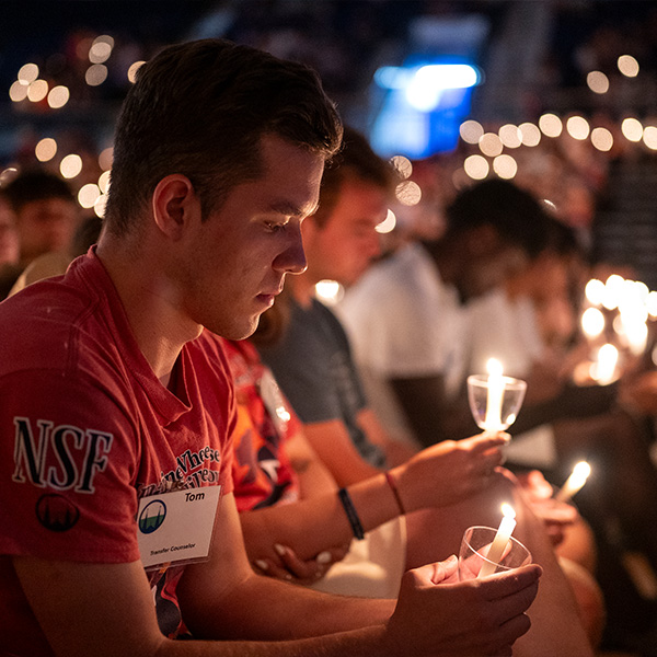 A male Orientation counselor sits holding a candle surrounded by other young people with candles. 